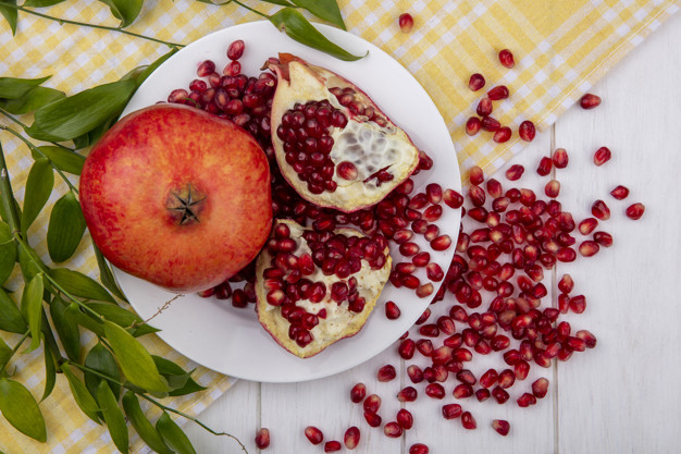 top view of whole pomegranate and pomegranate pieces with berries in plate and leaves on plaid cloth with pomegranate berries on wooden background